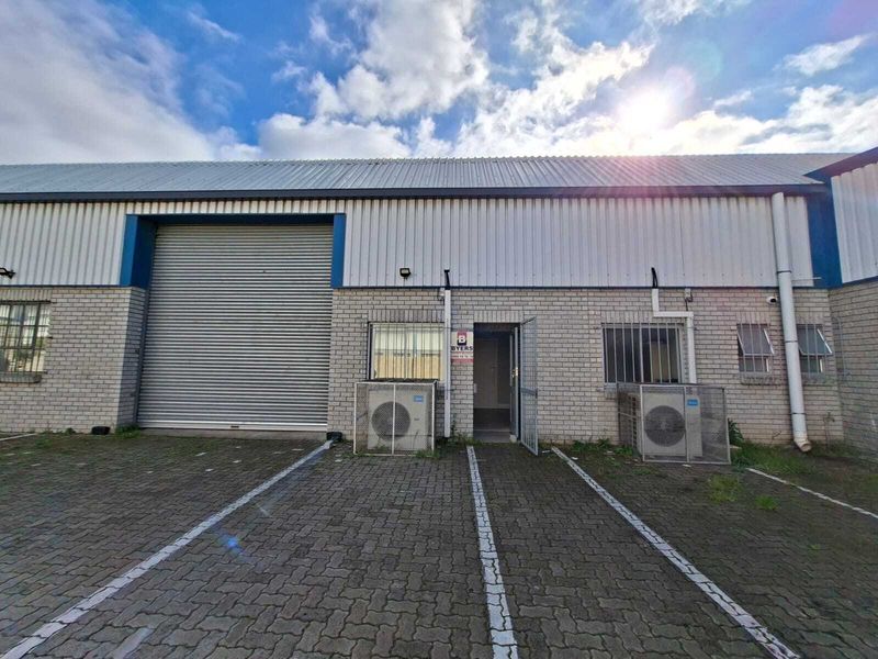 Industrial unit exterior featuring a large roller shutter door, security gate, and paved parking area with external air conditioning units.
