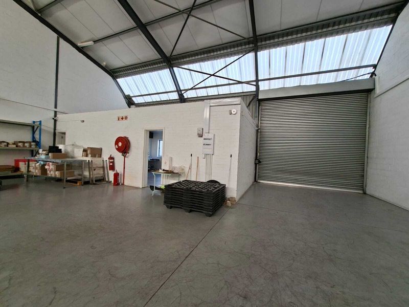 Spacious industrial warehouse unit featuring high ceilings with exposed steel trusses, translucent roof panels for natural light, and a large roller shutter door.
