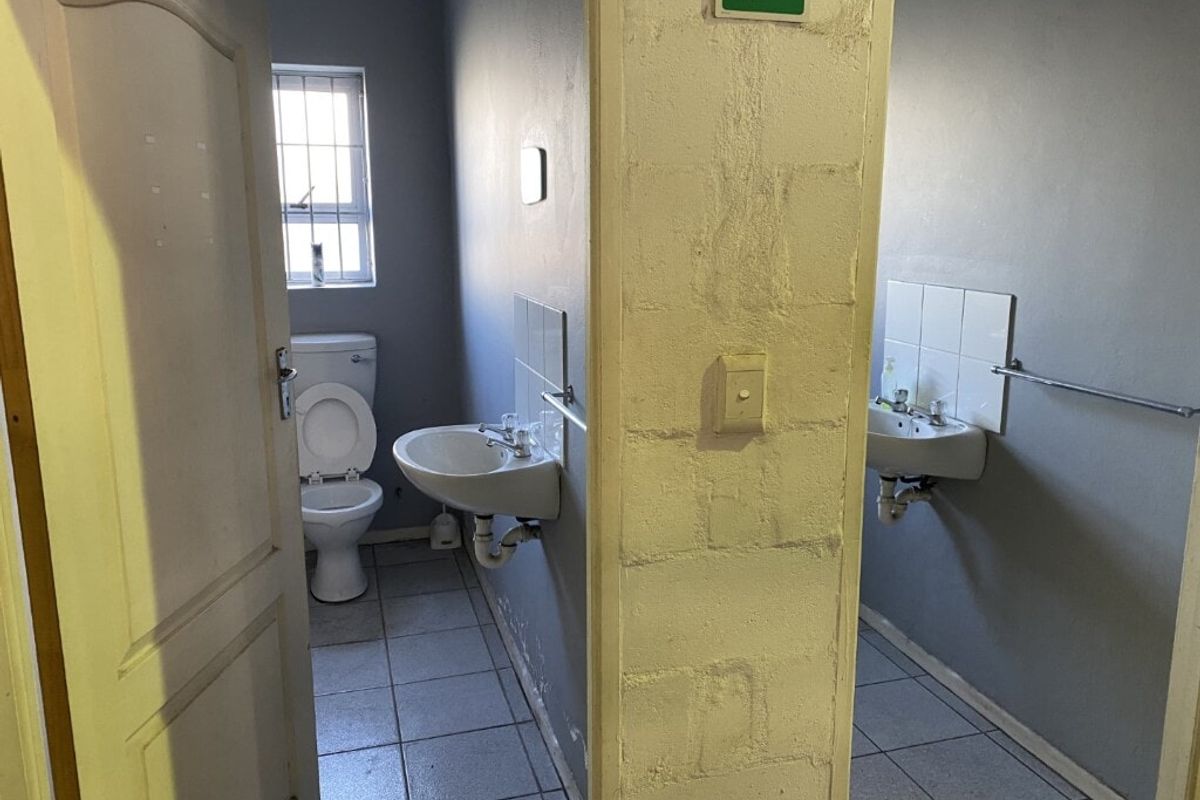 Functional industrial-style bathroom featuring dual washbasins, tiled flooring, and a private toilet cubicle with security-barred window.