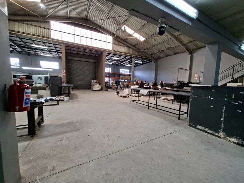 Spacious industrial warehouse area featuring high ceilings with exposed steel trusses, concrete flooring, and a large roller shutter door for easy access.