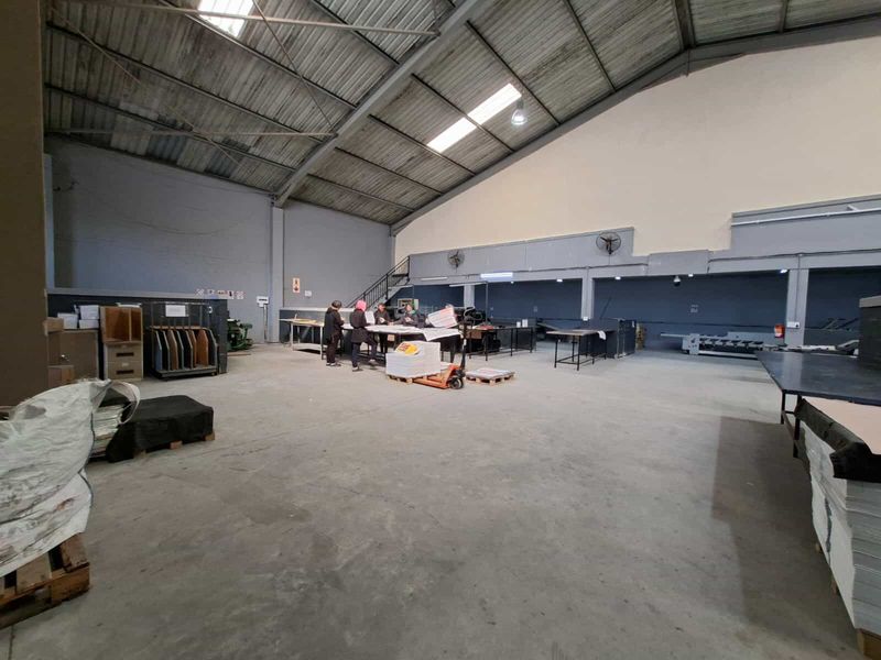 Spacious industrial warehouse area featuring high ceilings with exposed steel trusses, skylights, concrete flooring, and wall-mounted ventilation fans.