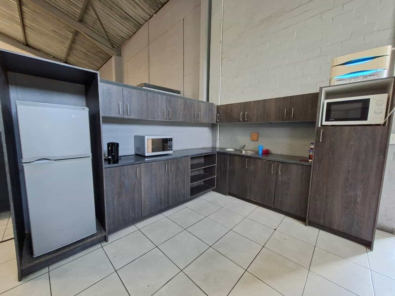 Industrial-style kitchenette featuring dark wood-grain cabinetry, laminate countertops, a stainless steel sink, and durable ceramic floor tiles under an exposed high ceiling.