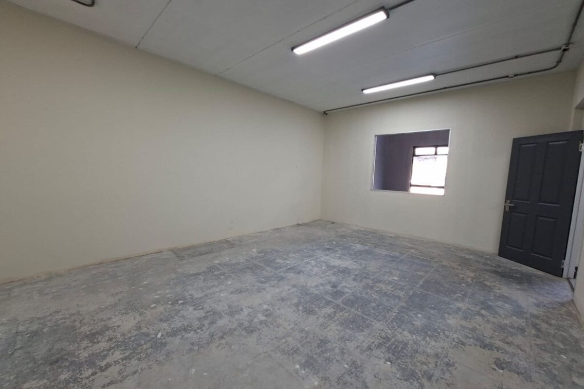 An empty industrial utility space featuring concrete flooring, exposed conduit lighting, and a solid core door, suitable for storage or workshop use.