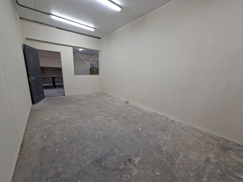 An empty industrial office space featuring unfinished concrete flooring, fluorescent lighting, and an internal window looking into an adjacent room.