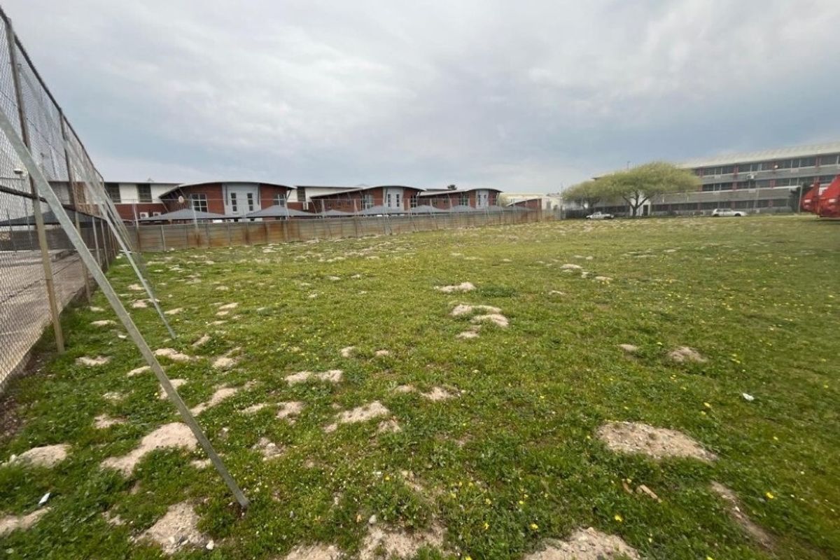 A view of an open, grassed industrial land plot enclosed by chain-link perimeter fencing, situated adjacent to existing commercial building structures.