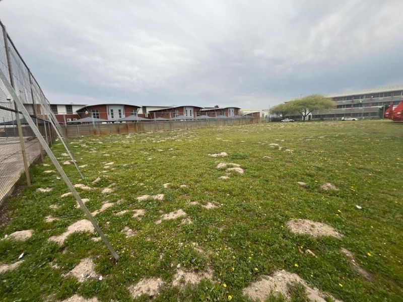 A view of an open, grassed industrial land plot enclosed by chain-link perimeter fencing, situated adjacent to existing commercial building structures.