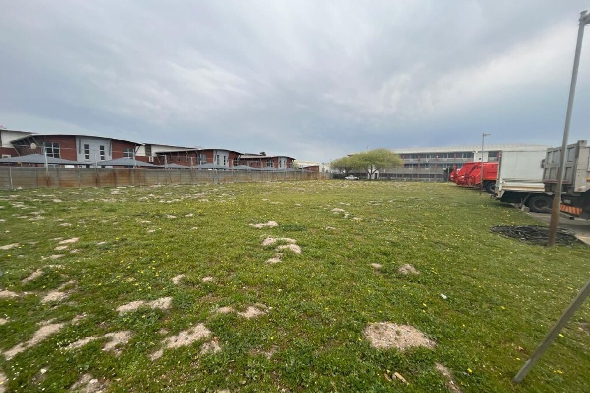 A view of an open industrial land plot featuring grassed terrain and perimeter fencing, adjacent to existing commercial building structures.