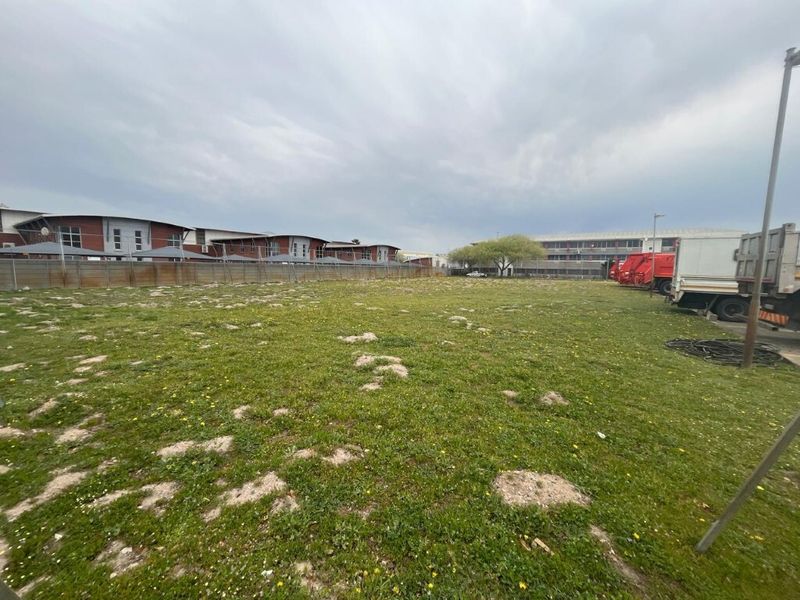 A view of an open industrial land plot featuring grassed terrain and perimeter fencing, adjacent to existing commercial building structures.