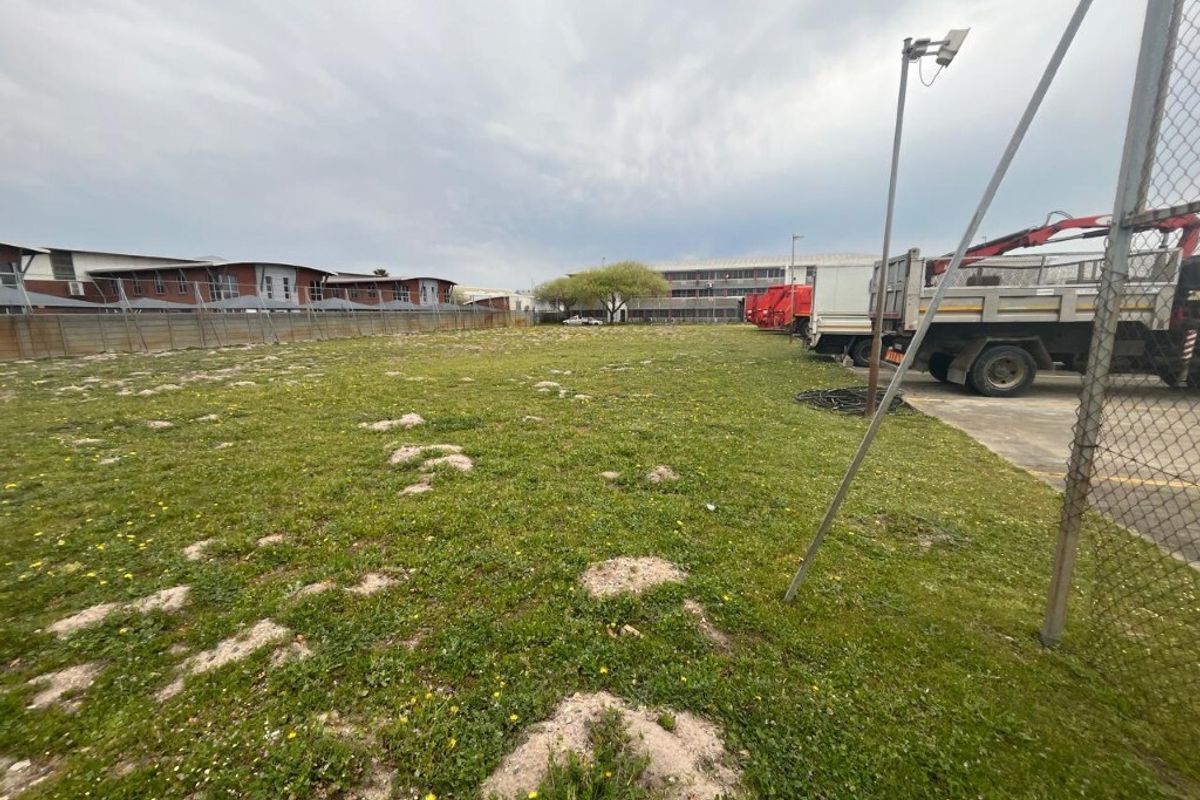 A vacant industrial land plot featuring a grassed area, perimeter chain-link fencing, and adjacent concrete hardstand with security lighting.