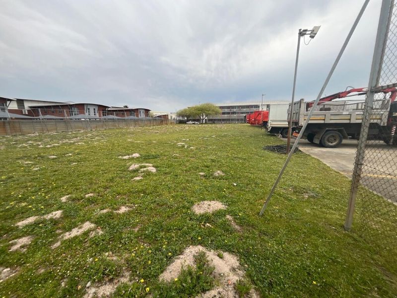 A vacant industrial land plot featuring a grassed area, perimeter chain-link fencing, and adjacent concrete hardstand with security lighting.