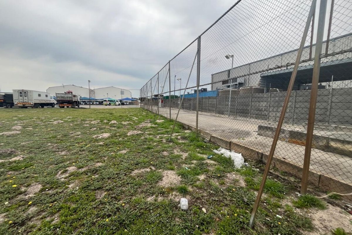 An industrial outdoor yard area featuring chain-link perimeter fencing, a concrete retaining wall, and a paved loading zone adjacent to warehouse structures.