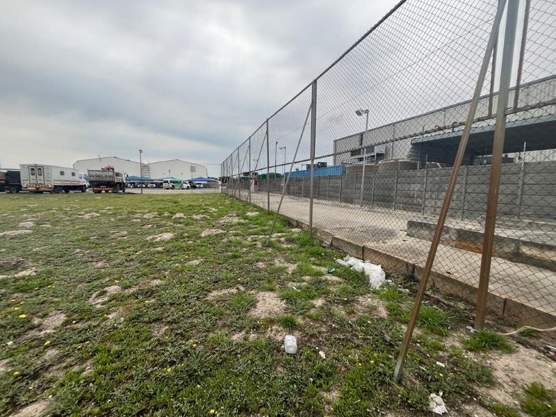 An industrial outdoor yard area featuring chain-link perimeter fencing, a concrete retaining wall, and a paved loading zone adjacent to warehouse structures.