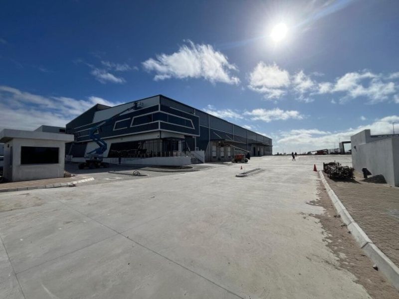 A modern industrial warehouse facility featuring a large concrete yard, security gatehouse, and multiple loading bays under a clear sky.