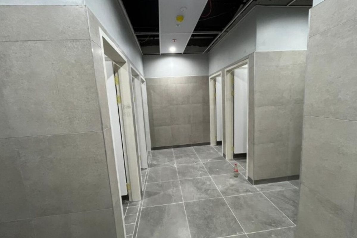 A commercial bathroom area under construction featuring grey tiled walls and flooring with an unfinished suspended ceiling and exposed infrastructure.