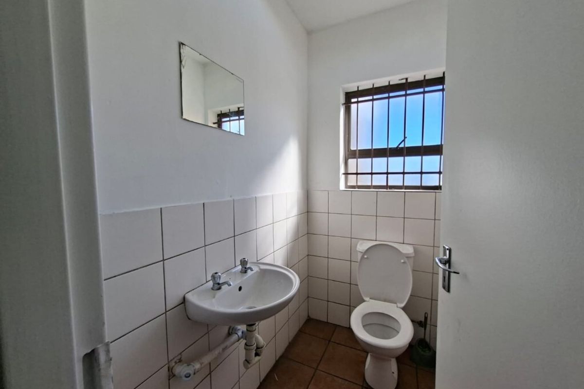 A functional industrial-style restroom featuring a wall-mounted basin, a standard toilet, and tiled wall finishes with a security-barred window.