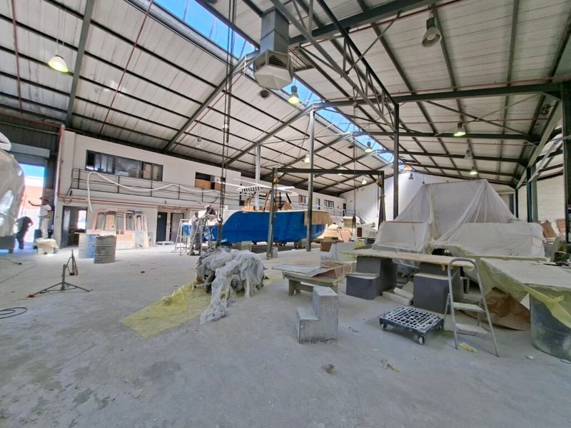 Spacious industrial workshop featuring high ceilings with exposed steel trusses, skylights, concrete flooring, and an overhead crane system, suitable for manufacturing or boat building.