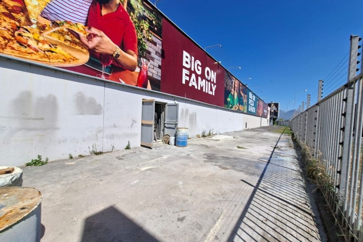 Rear exterior area of an industrial property featuring concrete paving, perimeter electric fencing, and a large wall-mounted advertising billboard.