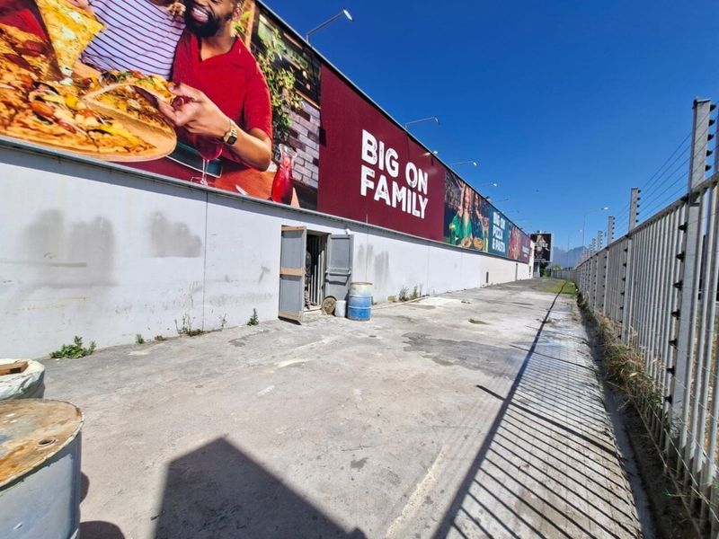 Rear exterior area of an industrial property featuring concrete paving, perimeter electric fencing, and a large wall-mounted advertising billboard.