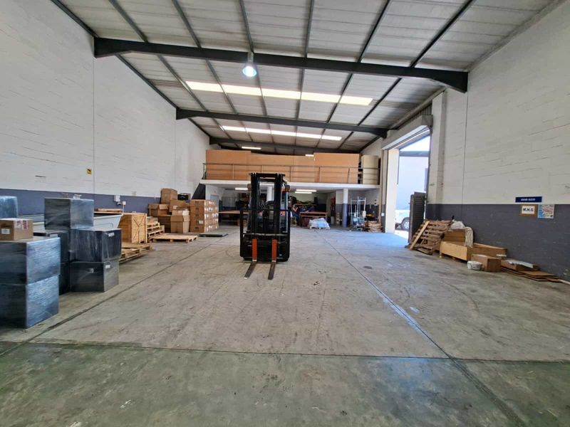 Spacious industrial warehouse unit featuring high ceilings with exposed steel trusses, a mezzanine level, concrete flooring, and a large roller shutter door for access.