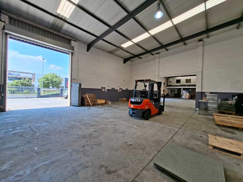 Spacious industrial warehouse unit featuring high ceilings, exposed steel trusses, concrete flooring, and a large roller shutter door for vehicle access.