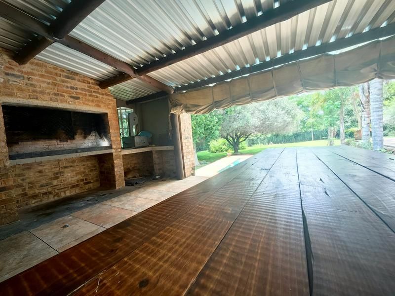 Covered outdoor patio featuring a built-in brick braai, exposed timber beams, and corrugated metal roofing, overlooking the garden and pool area.