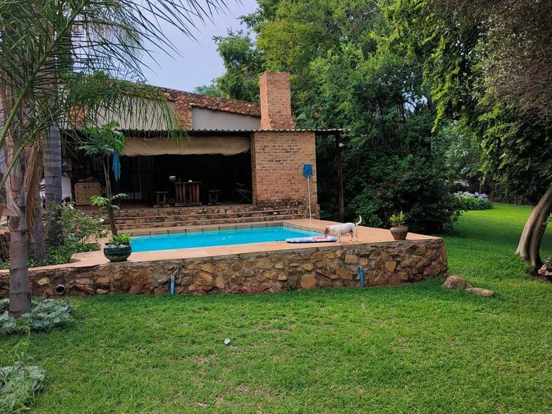 Outdoor entertainment area featuring a swimming pool with stone-clad retaining wall, adjacent covered patio with a brick fireplace, and established garden surroundings.