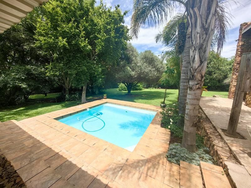 A well-maintained outdoor pool area featuring a tiled surround, stone-clad structural elements, and a covered patio overlooking a lush, established garden.