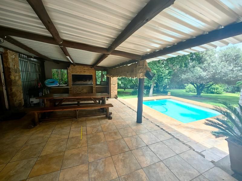 Covered outdoor patio featuring a built-in braai, tiled flooring, and timber beams, overlooking a private swimming pool and garden area.