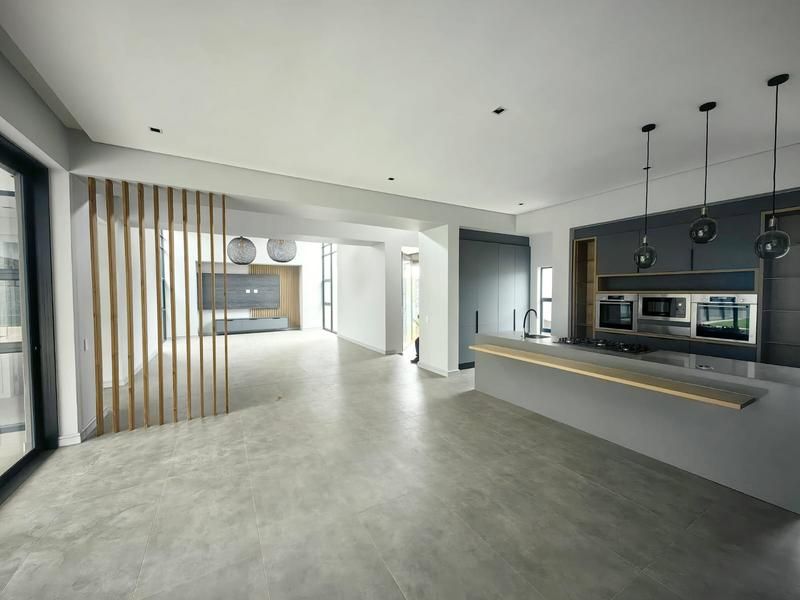 Spacious open-plan living and kitchen area featuring modern grey cabinetry, integrated appliances, a timber slat divider, and large format floor tiles.