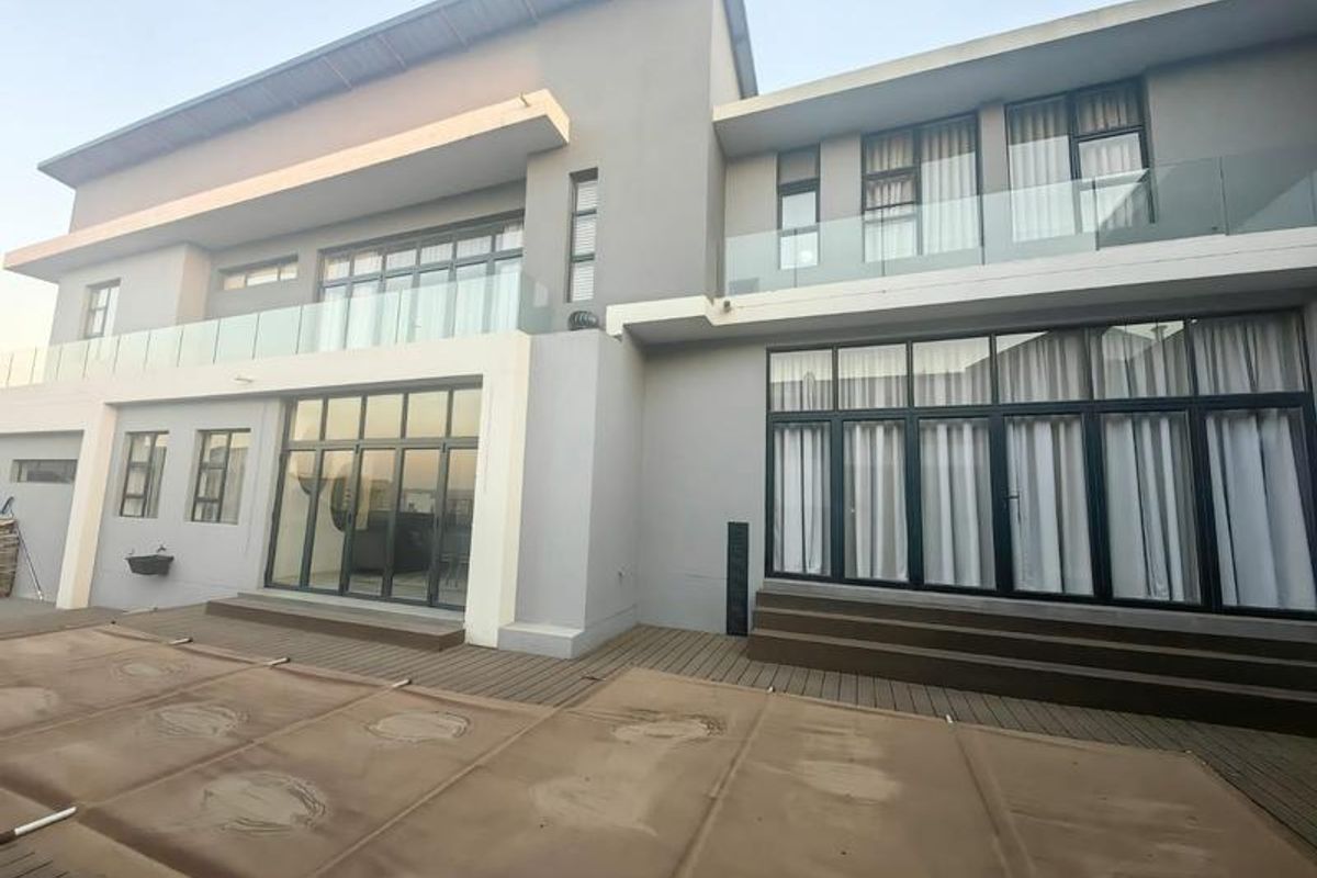 Rear elevation of a modern double-story house featuring extensive glass sliding doors, upper-level balconies with glass balustrades, and a timber deck with a pool cover.