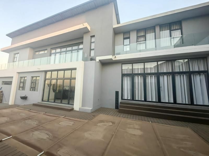 Rear elevation of a modern double-story house featuring extensive glass sliding doors, upper-level balconies with glass balustrades, and a timber deck with a pool cover.