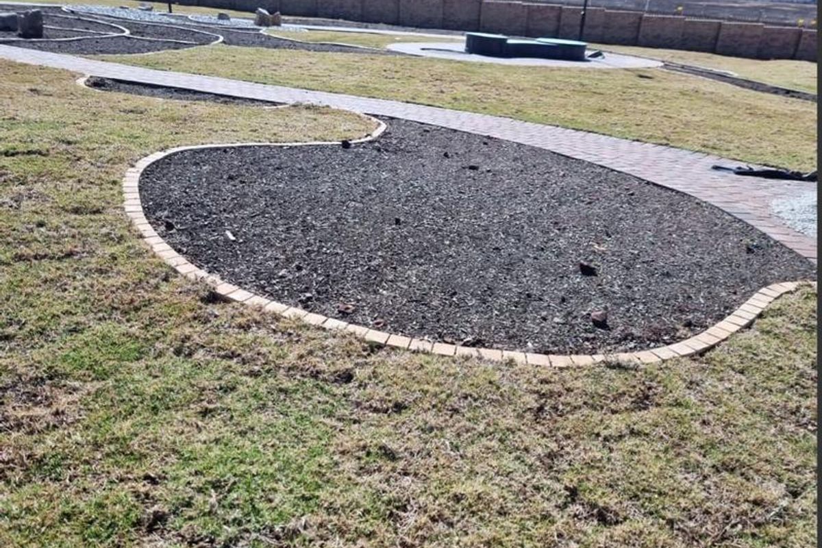 A landscaped garden area featuring a paved walkway, brick-edged mulched garden beds, and a perimeter boundary wall.