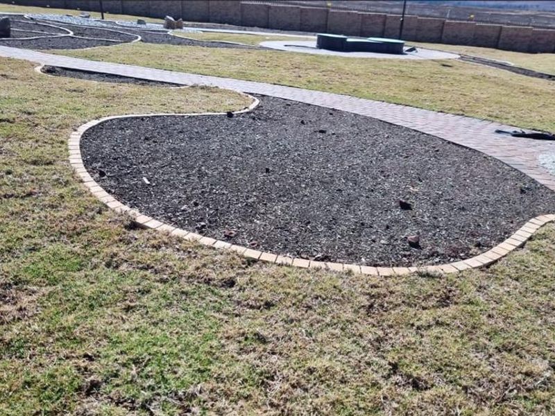 A landscaped garden area featuring a paved walkway, brick-edged mulched garden beds, and a perimeter boundary wall.