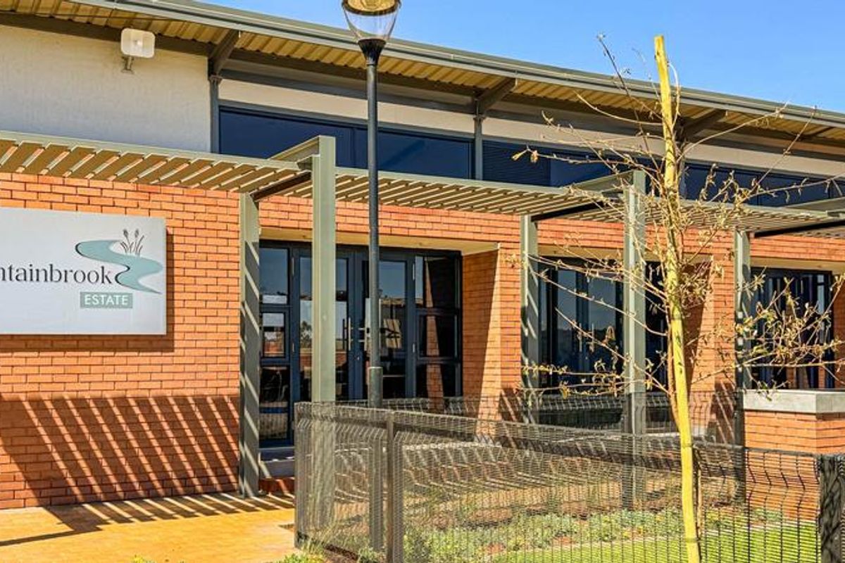 Modern brick clubhouse building featuring a metal pergola, double glass entrance doors, and estate signage, set within a well-maintained landscaped area.