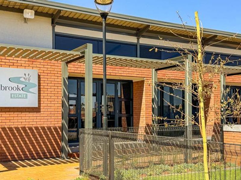Modern brick clubhouse building featuring a metal pergola, double glass entrance doors, and estate signage, set within a well-maintained landscaped area.