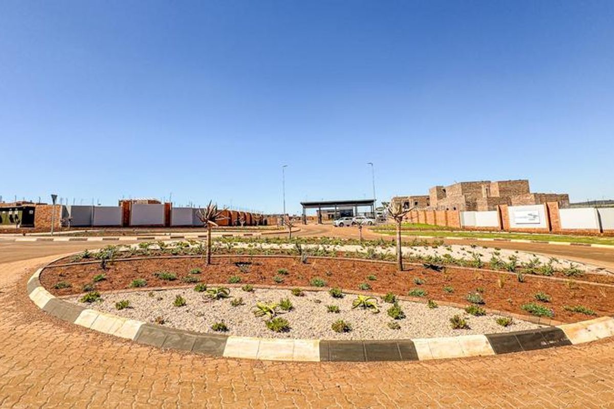 A view of the estate entrance featuring a landscaped roundabout, paved roads, and perimeter walling leading to the security gatehouse.