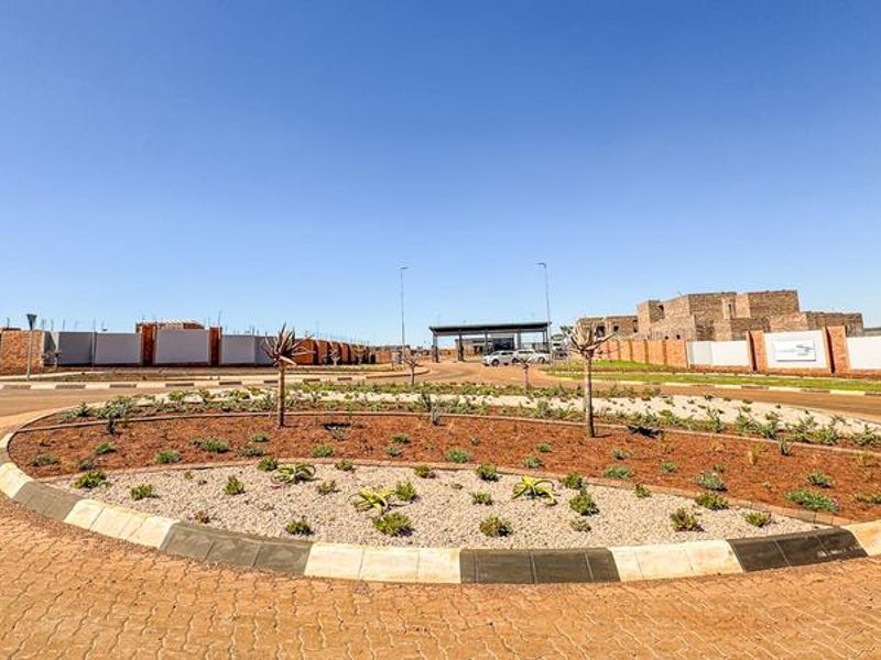 A view of the estate entrance featuring a landscaped roundabout, paved roads, and perimeter walling leading to the security gatehouse.