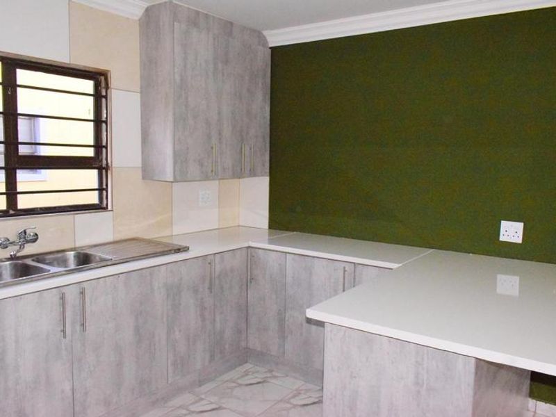 A clean, modern kitchen featuring light grey wood-grain cabinetry, white stone countertops, a stainless steel sink, and a contrasting dark green feature wall.