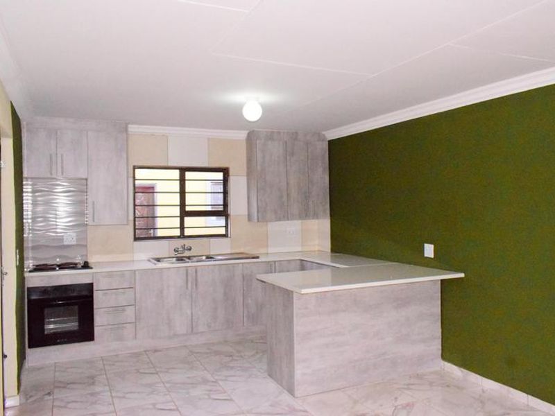 A clean, modern kitchen featuring light wood-grain cabinetry, stone countertops, built-in oven, hob, and tiled flooring with a contrasting green feature wall.