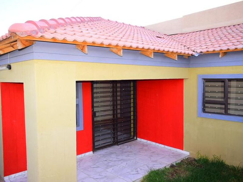 A covered patio area featuring tiled flooring, a security gate, and a tiled roof, requiring some exterior maintenance and paint touch-ups.