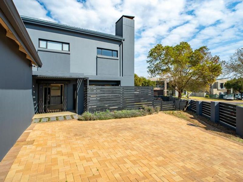 Modern double-story residence featuring a paved driveway, contemporary grey facade, chimney stack, and secure slatted fencing along the property boundary.