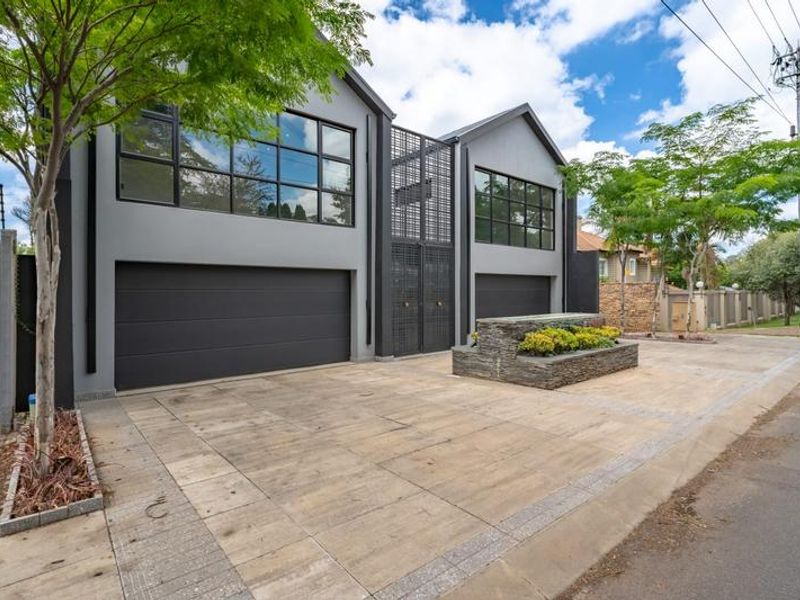 Modern double-story facade featuring dark grey exterior finish, large industrial windows, double garage doors, and a stone-clad planter box in the paved driveway.