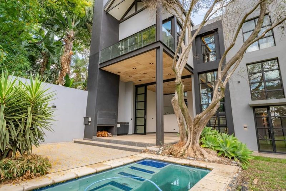 Modern rear exterior featuring a covered patio with built-in fireplace, glass-railed balcony, and a swimming pool set within a landscaped garden.
