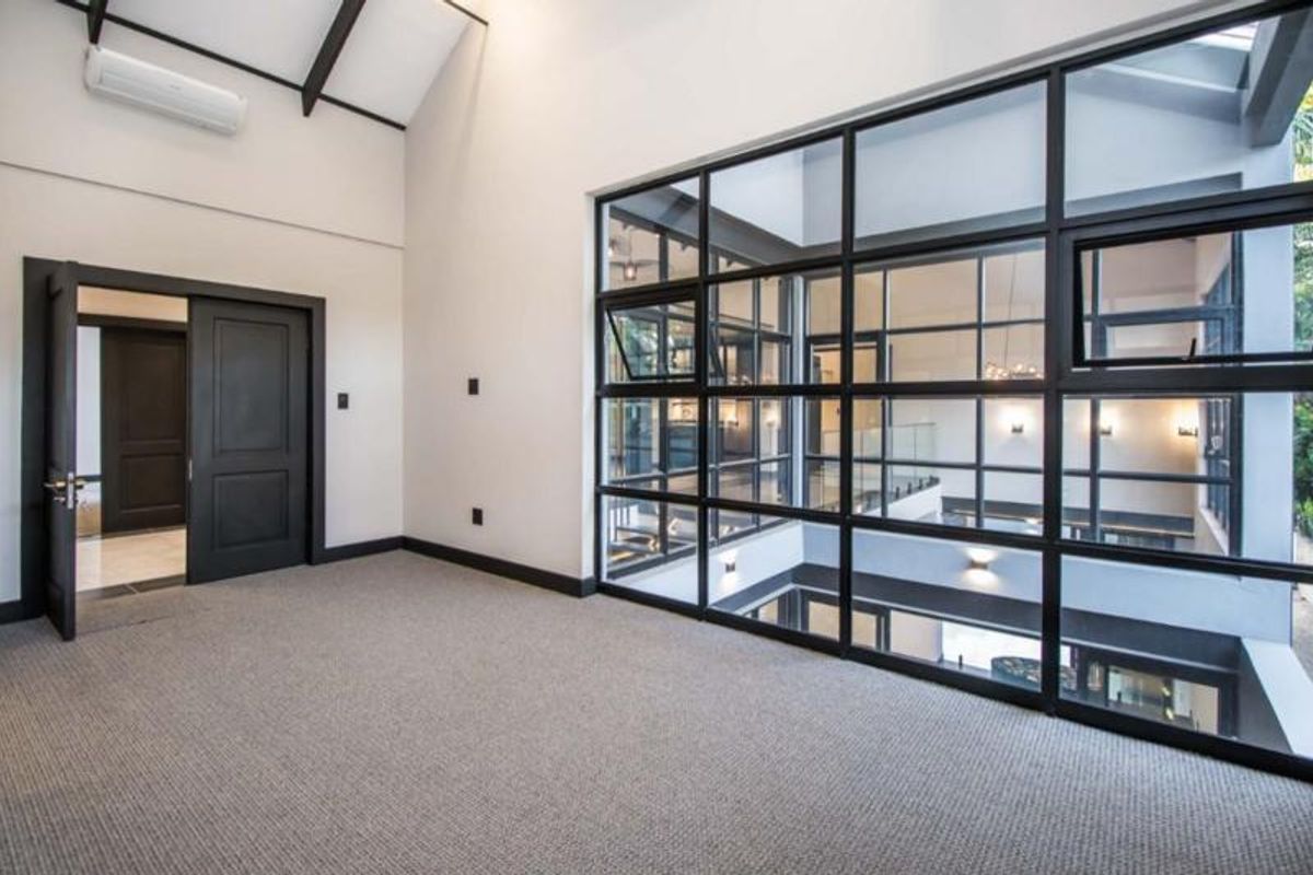 Spacious bedroom featuring a vaulted ceiling with exposed beams, carpeted flooring, and a large black-framed glass partition overlooking the interior space.