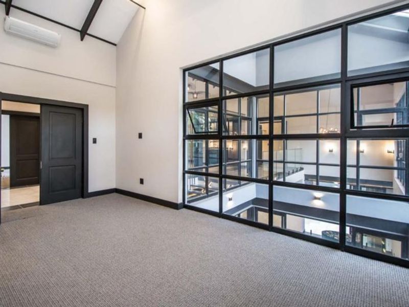 Spacious bedroom featuring a vaulted ceiling with exposed beams, carpeted flooring, and a large black-framed glass partition overlooking the interior space.