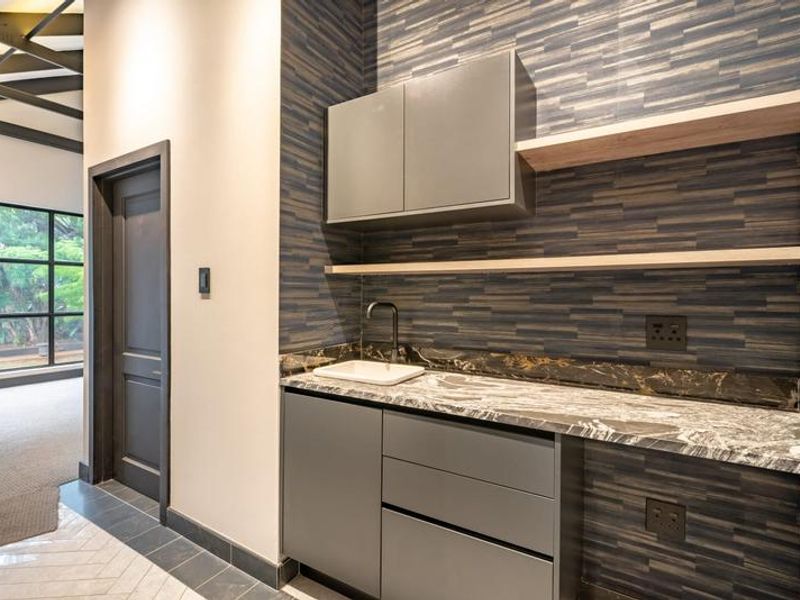 A modern, well-appointed scullery featuring sleek grey cabinetry, a striking stone countertop, undermount sink, and stylish floating shelves against a textured feature wall.