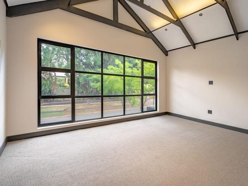 Spacious bedroom featuring a vaulted ceiling with exposed timber beams, large black-framed windows, and neutral carpeting, offering a bright and airy atmosphere.