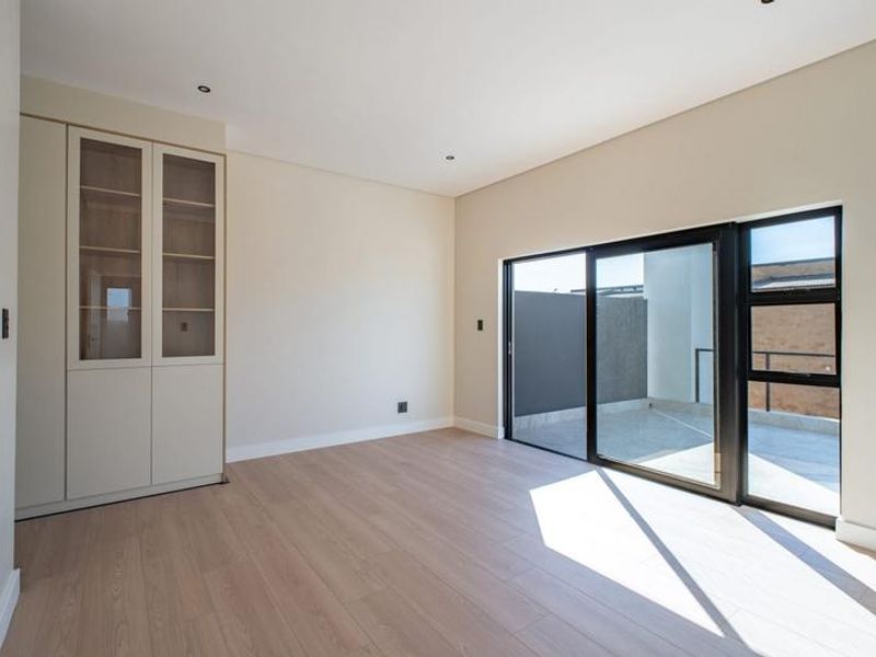 A bright, modern bedroom featuring light laminate flooring, built-in cabinetry, and a large sliding glass door providing access to an outdoor balcony.