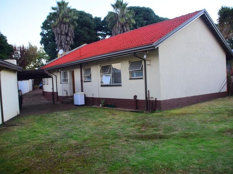 Rear view of a single-story house featuring a red tiled roof, plastered walls, and an attached carport area with a surrounding lawn.