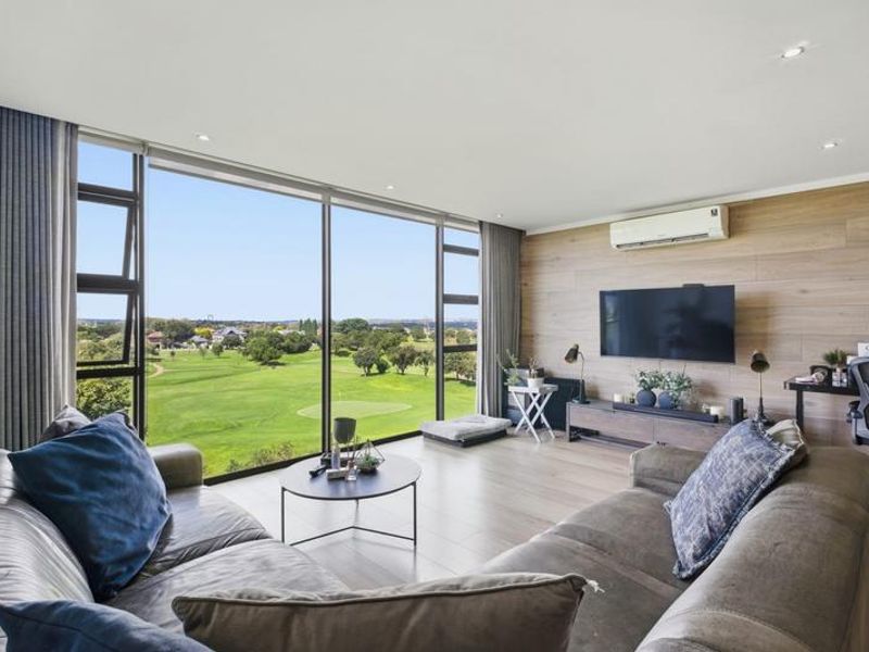 A bright, modern living room featuring floor-to-ceiling windows with expansive golf course views, timber-look wall paneling, and integrated air conditioning.